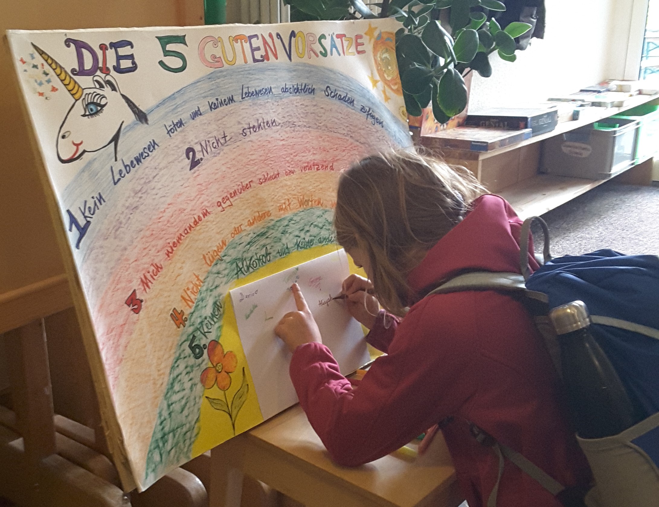 A child writes on a sheet of paper in front of a colorful poster titled ‘The Five Good Precepts’
