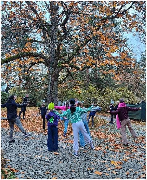 Children and helpers stand in a circle around a tree and do morning exercise together